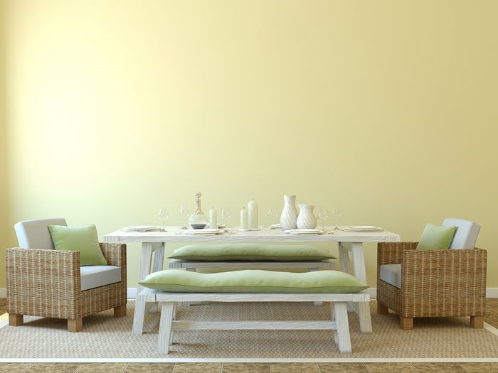 A white picnic table and matching benches set with elegant, white flatware and candles in a yellow room. Wicker chairs sit on either side of the table.